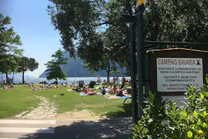 Campingwiese am See mit vielen Liegen, Bäumen und Blick auf Berge, im Vordergrund ein Schild „Camping Bavaria“.