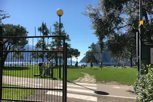 Campingplatz am See: grüner Rasen, Bänke und Liegen, im Hintergrund Berge und Wasser unter blauem Himmel.