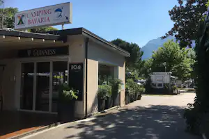 Schild „Camping Bavaria“ am Eingang, Rezeption und Gebäude mit Glasfront, Pflanzen und Bäume am Weg.