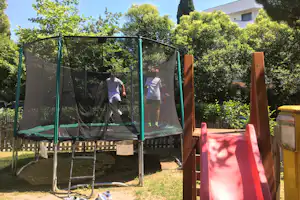 Trampolin im Campinggarten mit grünem Netz, daneben Rutsche und Bäume vor einem weißen Gebäude.