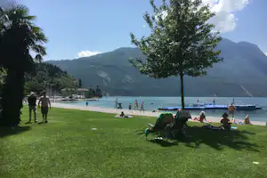 Grüner Campingplatz am See mit Baum im Vordergrund, Liegewiese, Steg und Blick auf die Berge.