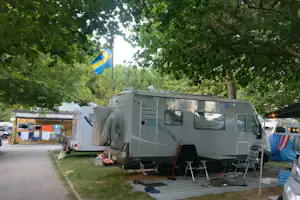 Wohnmobil auf dem Stellplatz im Grünen, mit Markise, Tisch und Stühlen unter Bäumen und Flagge.
