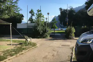 Campingweg zum See mit Rasenfläche, Hecken und Laternen, im Hintergrund Berge und Bäume.