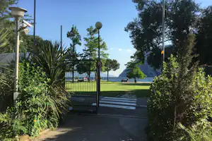 Campingzaun und Rasenfläche am Seeufer, Bäume und Laternen, im Hintergrund Menschen am Strandweg.