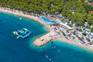 Luftaufnahme einer Badebucht mit Sandstrand, türkisfarbenem Wasser, Schwimmbecken und vielen Liegen am Ufer.