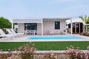 Moderner Bungalow mit Terrasse, zwei Stühlen und Tisch, Pool im Vordergrund und Liegen auf dem Campingplatz.