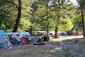 Campingplatz mit Wohnwagen und Zelten am Stellplatz, davor Liegestühle und ein Schlauchboot, umgeben von Bäumen.