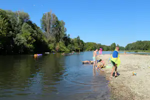 Badegäste am Seeufer mit Kiesstrand, Schwimmreifen im Wasser und Bäumen am gegenüberliegenden Ufer.