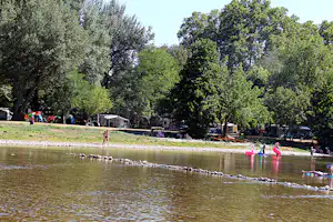 Campingplatz am See mit Olivenbäumen, Liegestühlen am Ufer und mehreren Personen im flachen Wasser.