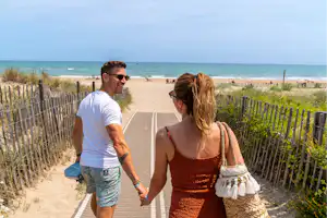 Paar geht auf einem Strandweg durch die Dünen, mit Blick auf das Meer und den Sandstrand.