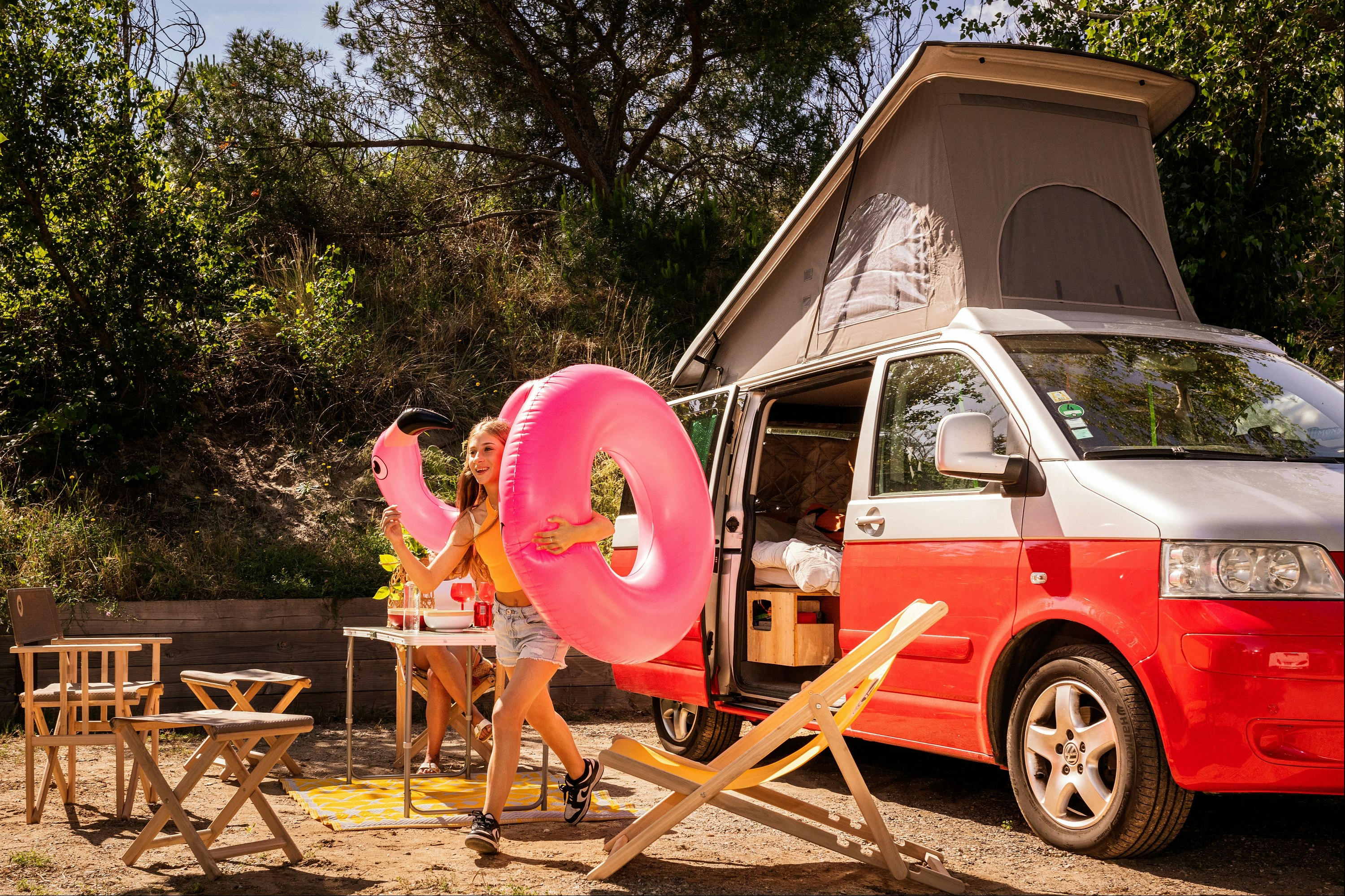 Roter Campingvan mit aufgestelltem Zelt, Tisch und Stühlen am Stellplatz im Grünen, daneben ein pinkfarbener Schwimmring.