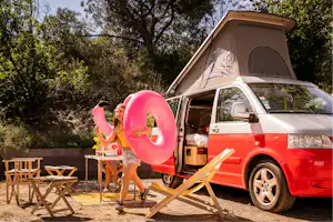 Roter Campingvan mit aufgestelltem Zelt, Tisch und Stühlen am Stellplatz im Grünen, daneben ein pinkfarbener Schwimmring.