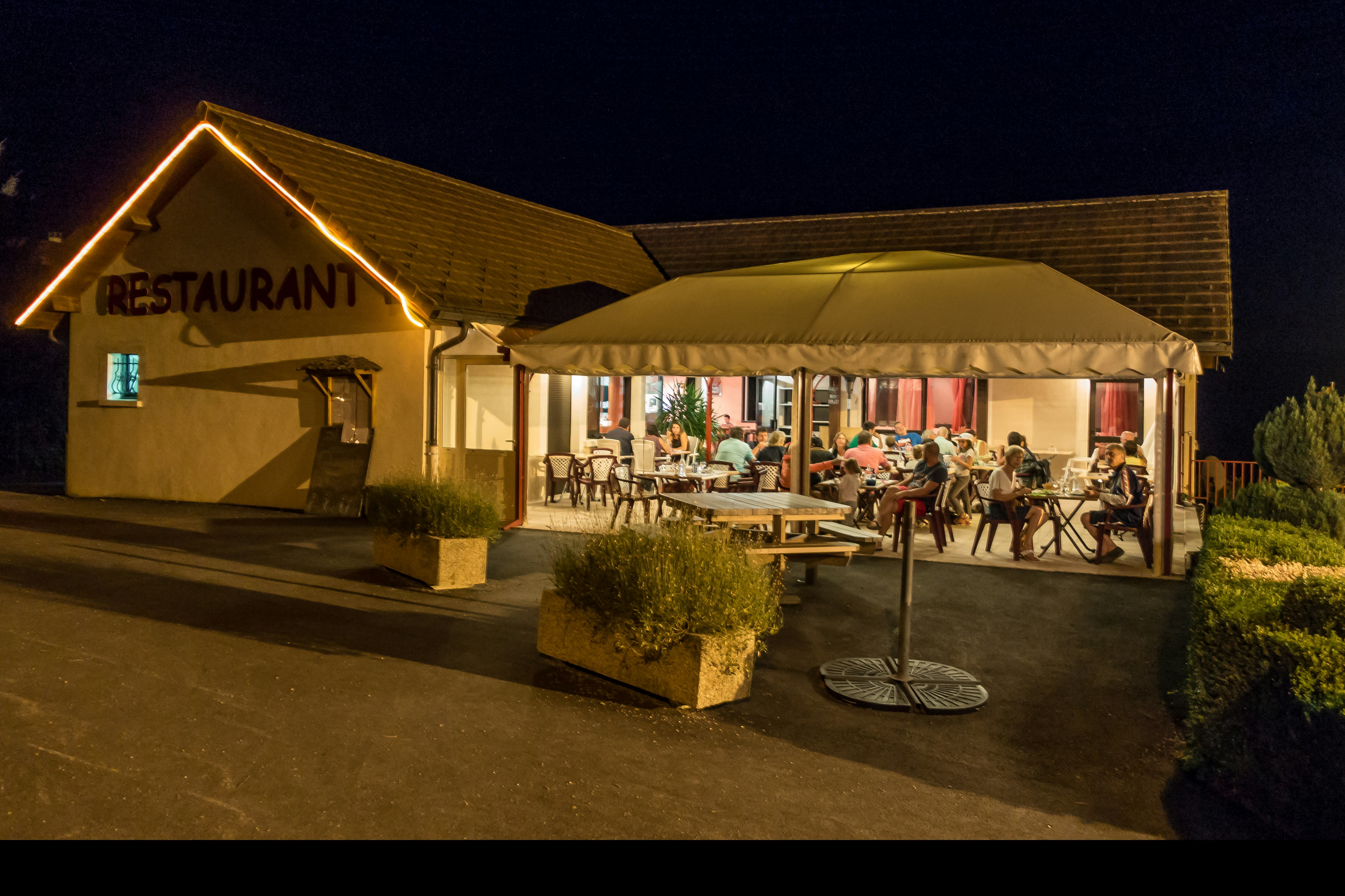Restaurant am Campingplatz bei Nacht, beleuchteter Eingang, Terrasse mit Tischen und Stühlen unter einem großen Sonnenschirm.