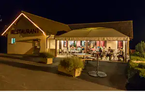Restaurant am Campingplatz bei Nacht, beleuchteter Eingang, Terrasse mit Tischen und Stühlen unter einem großen Sonnenschirm.