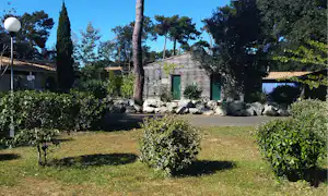 Gepflegter Campingplatz mit Rasen, Hecken und mehreren Bäumen vor einem Gebäude mit grünen Türen und Steinmauer.