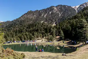 Campingplatz am See im Tal: viele Autos, Picknicktische und Menschen am Ufer vor bewaldeten Bergen.