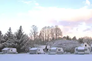 Wohnwagen und Zelte auf einem Campingplatz im Schnee, umgeben von verschneiten Bäumen und einem Winterhimmel.