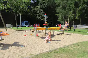 Sandspielplatz mit gelbem Spielgerät, Bank und Zelt im Hintergrund zwischen Bäumen auf dem Campinggelände.
