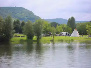 Zelt und Wohnmobile am Seeufer auf einer Campingwiese, umgeben von Bäumen und Hügeln im Hintergrund.