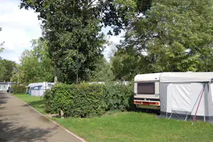 Wohnwagen und Zelt auf einem Campingplatz, umgeben von Hecken und großen Bäumen entlang eines gepflasterten Wegs.