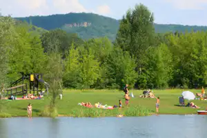 Campingplatz am See mit Spielbereich, Liegestühlen und Sonnenschirm, im Hintergrund grüne Bäume und Hügel.