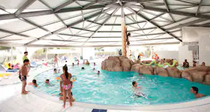 Überdachter Innenpool auf einem Campingplatz, Menschen im Wasser, große Glaswände und helle Fliesen im Beckenbereich.