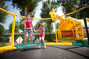 Kinder auf dem Trampolin-Spielplatz im Campingbereich, mit gelbem Netz und Rutsche, im Hintergrund Bäume und Zaun.