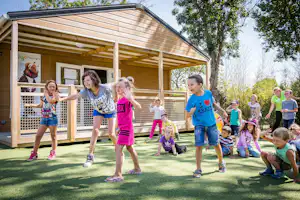 Kinder spielen vor einem Holz-Spielhaus am Campingplatz, mit Rasenfläche, Zaun und Bäumen im Hintergrund.