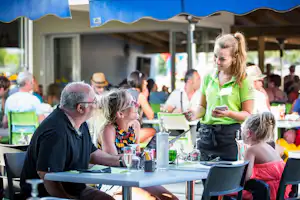 Familie am Tisch im Campingrestaurant, Kinder und Erwachsene, gedeckte Tische mit Gläsern und Speisen unter Sonnenschirmen.