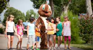 Ein großes Maskottchen steht auf dem Weg, Kinder und Erwachsene laufen im Campingpark zwischen Bäumen.