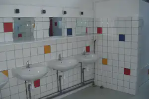 Zwei Waschbecken mit Armaturen in einem gefliesten Sanitärraum, dahinter Spender und farbige Fliesenfelder.