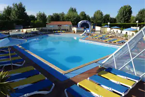 Schwimmbad mit blauem Wasser, Liegen und Sonnenschirmen am Campingplatz, im Hintergrund Gebäude und Bäume.