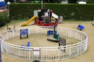 Spielplatz mit Kletterturm und gelber Rutsche auf Sandfläche, umgeben von Hecken und einem weißen Metallzaun.