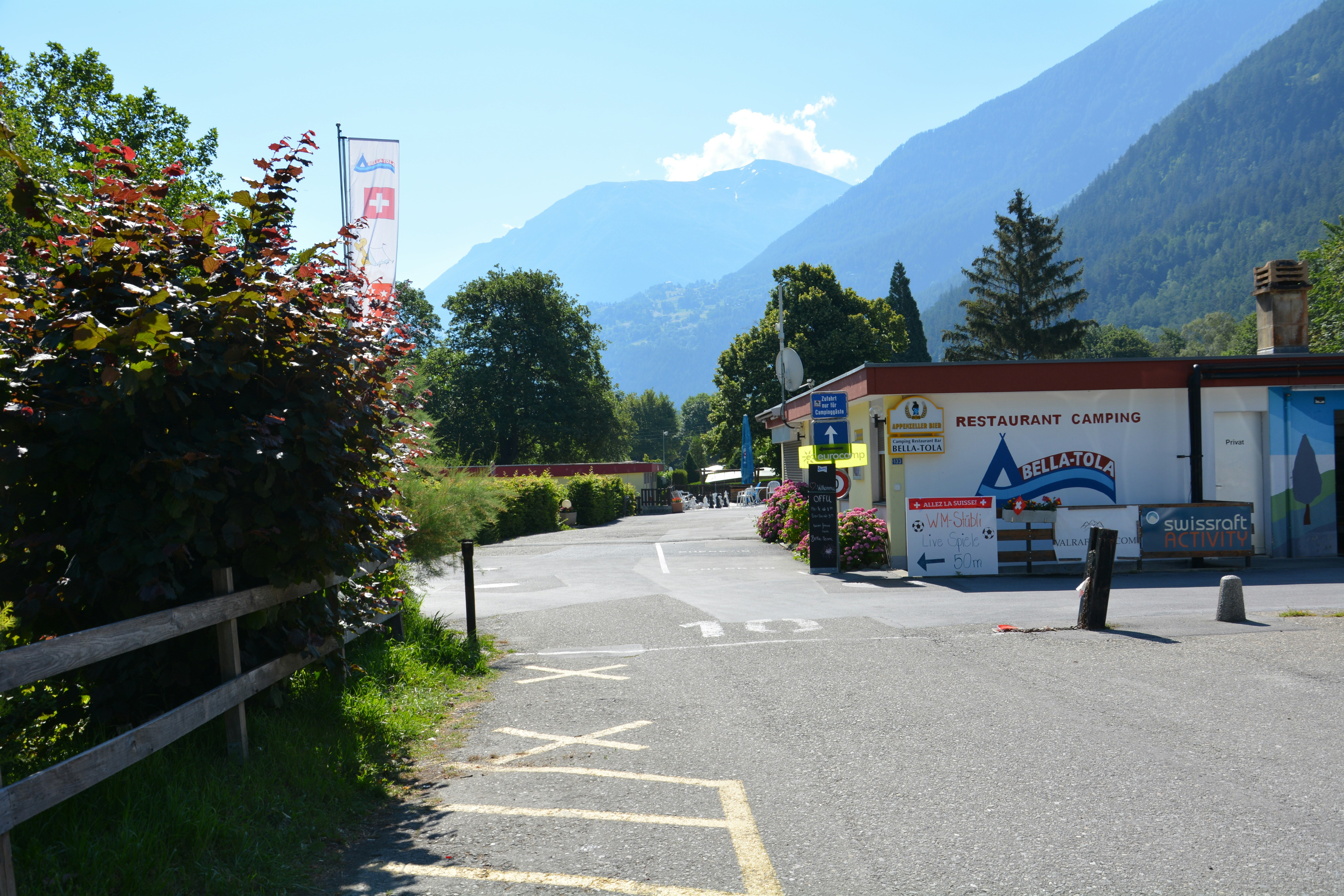 Eingang zum Restaurant Camping Bella-Tola am Weg, mit Fahnen, Blumenbeeten und Blick auf grüne Berge.