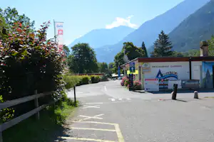 Eingang zum Restaurant Camping Bella-Tola am Weg, mit Fahnen, Blumenbeeten und Blick auf grüne Berge.