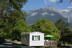 Weißes Mobilheim mit Terrasse und grüner Markise, umgeben von Bäumen, Blick auf die Alpen im Hintergrund.