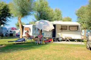 Wohnwagen mit Vorzelt und Markise auf dem Campingplatz, zwei Personen sitzen auf Klappstühlen im Schatten eines Baums.