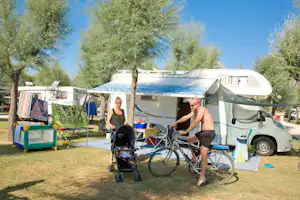 Wohnwagen auf dem Campingplatz unter Bäumen, mit Markise, Fahrrädern und Campingstühlen auf dem Stellplatz.