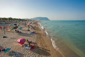 Strand mit vielen Liegestühlen und bunten Sonnenschirmen, Menschen im Wasser und Blick auf das Meer und einen Hügel.