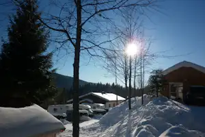 Winterlicher Campingplatz mit verschneiten Wohnwagen, Bäumen und Hütten, Blick in die Berge bei tiefem Himmel.