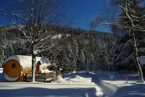 Winterlicher Campingplatz: Fasssauna mit Holzfassade, verschneiter Terrasse und Zaun, im Hintergrund Nadelwald und blauer Himmel.
