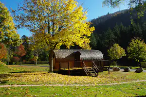 Holzhütte auf Campingplatz mit Holzterrasse, umgeben von bunten Bäumen und Hecken im Tal.