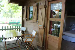 Holzunterkunft mit Terrasse, Tisch und Stühlen, große Fensterfront und Blick in den Garten mit Bäumen.