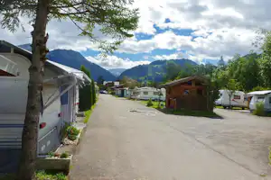 Reihe von Wohnwagen auf einem Campingplatz, gepflasterter Weg, Hecken und Bäume, im Hintergrund Berge und Wolken.