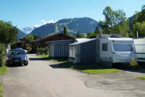 Stellplatz mit Wohnwagen und Auto auf dem Weg, im Hintergrund Berge und Bäume vor einem Campinggebäude.
