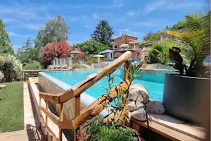 Holzstühle auf einer Terrasse am Campingplatz neben einem türkisfarbenen Pool, umgeben von Bäumen und Gartenwegen.