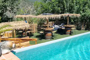 Schwimmbecken mit türkisfarbenem Wasser, Holzterrasse und Liegestühlen, im Hintergrund Bäume und ein Grillbereich.