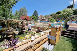 Blick auf einen Campinggarten mit Holzterrasse, Sitzbank und Blumenbeeten, im Hintergrund ein Pool und Gebäude.