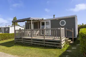 Wohnwagen mit Holzveranda und Geländer, davor Rasen und ein Weg, im Hintergrund weitere Wohnwagen im Campingbereich.