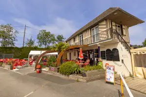 Campinghaus mit Balkon und Terrasse, davor Gehweg und Sitzbereich, im Hintergrund Bäume und ein Parkplatz.